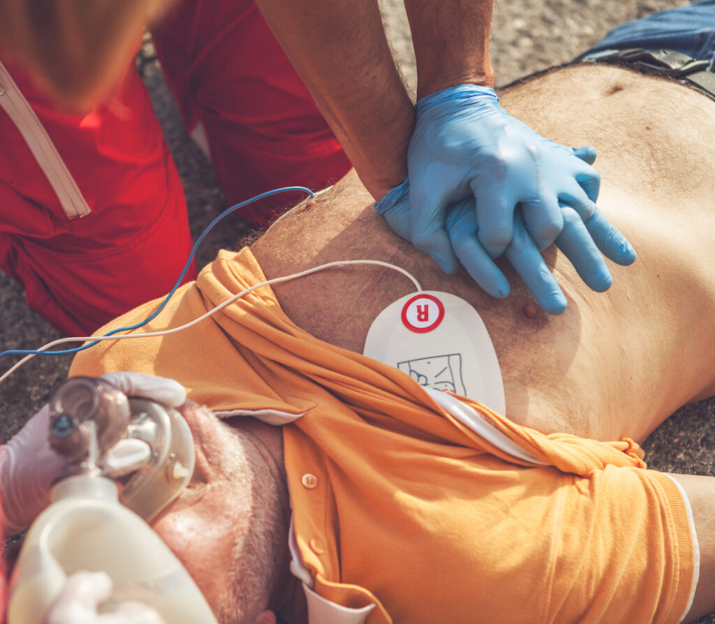 Ein Mann mit orangenem T-Shirt wird mit Defibrillator und einem Sanitäter mit blauen Handschuhen reanimiert.